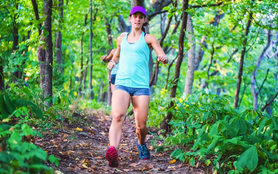 A woman runs down a green leafy path in spring wearing a TrailHeads hat