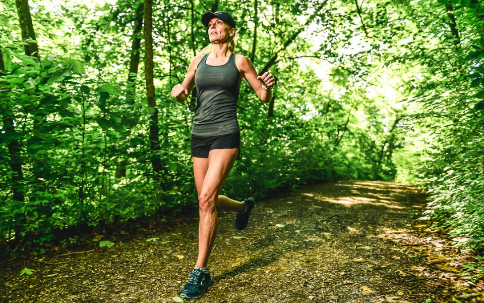 A woman runs along a sunny trail wearing a TrailHeads UV Hat